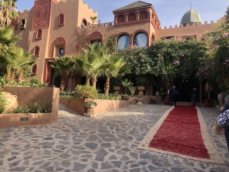 The entrance courtyard of Kasbah Tamadot, with carved pink-ochre walls, palms, flowering shrubs and a red rug laid across the cobbles to the door
