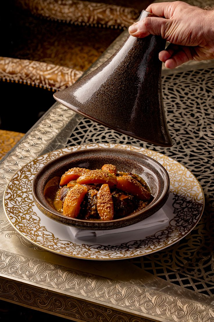 A hand lifting the lid from a small tagine of lamb and sesame served on a gold-rimmed plate