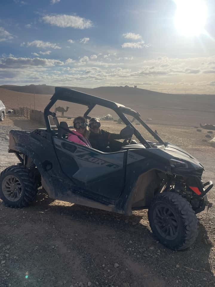 Two riders in helmets and goggles sitting in a two-seater desert buggy in the Agafay desert near Marrakech, with a camel on the ridge behind