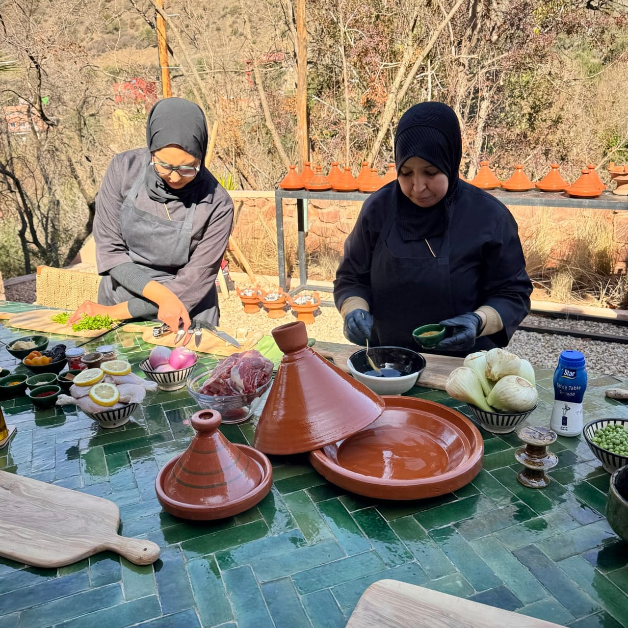 Two Moroccan cooks in aprons chopping herbs and measuring spices at a green tiled table with tagine dishes and ingredients