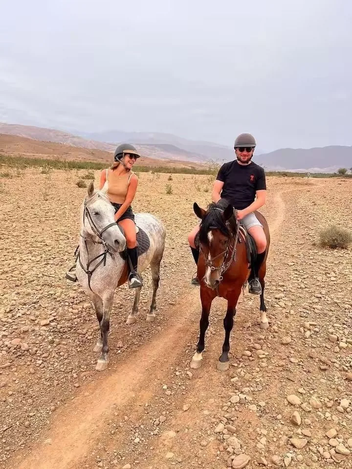 Dos jinetes con casco montando caballos en un camino del desierto de Agafay con colinas secas de fondo