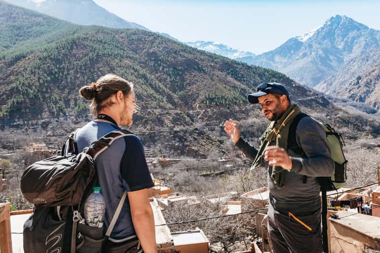 A mountain guide talking with a trekker over glasses of tea above an Imlil valley village with snow-capped peaks behind