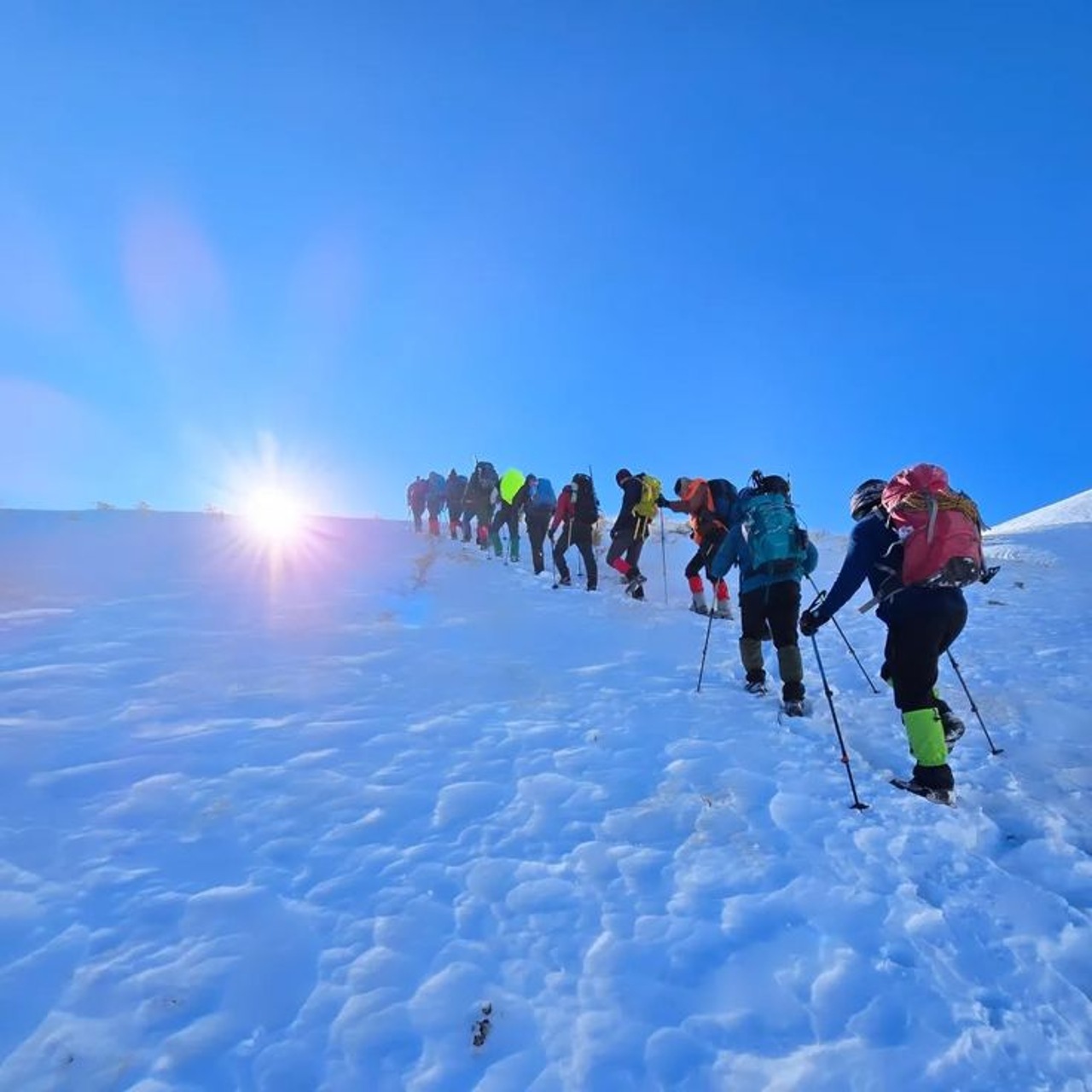 A line of trekkers ascending a snow slope towards the sun on the High Atlas