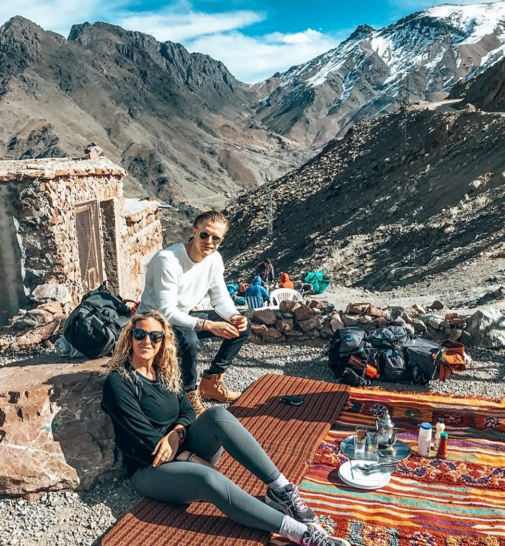 Two guests resting on Berber rugs with a mint tea tray at a stone shelter below snowy peaks