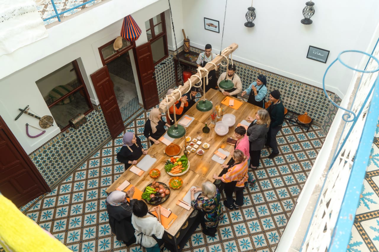 Overhead view of a cooking-class group gathered around a long wooden preparation table in a tiled Marrakech riad