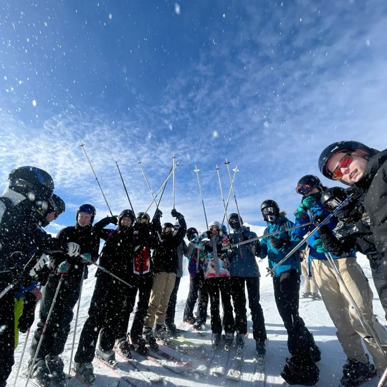 A large group of skiers raising their poles together on a snowy slope as snow blows around them