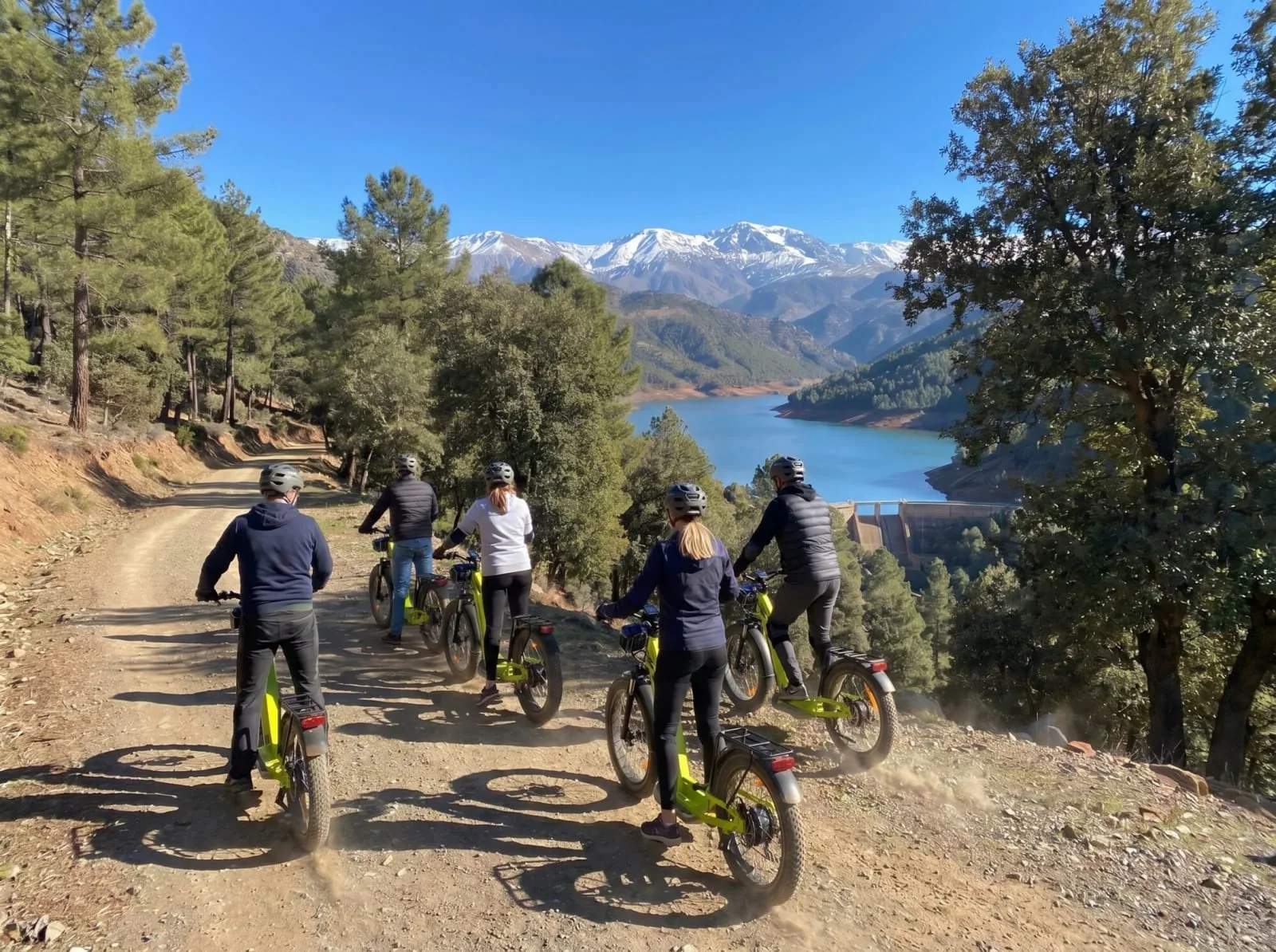 Riders in helmets descending a forest track on e-scooters above an Atlas mountain lake