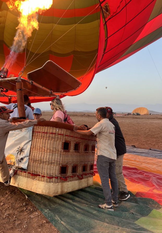 Ground crew steadying a wicker hot-air balloon basket with passengers aboard as the burner fires at sunrise on the Marrakech plain