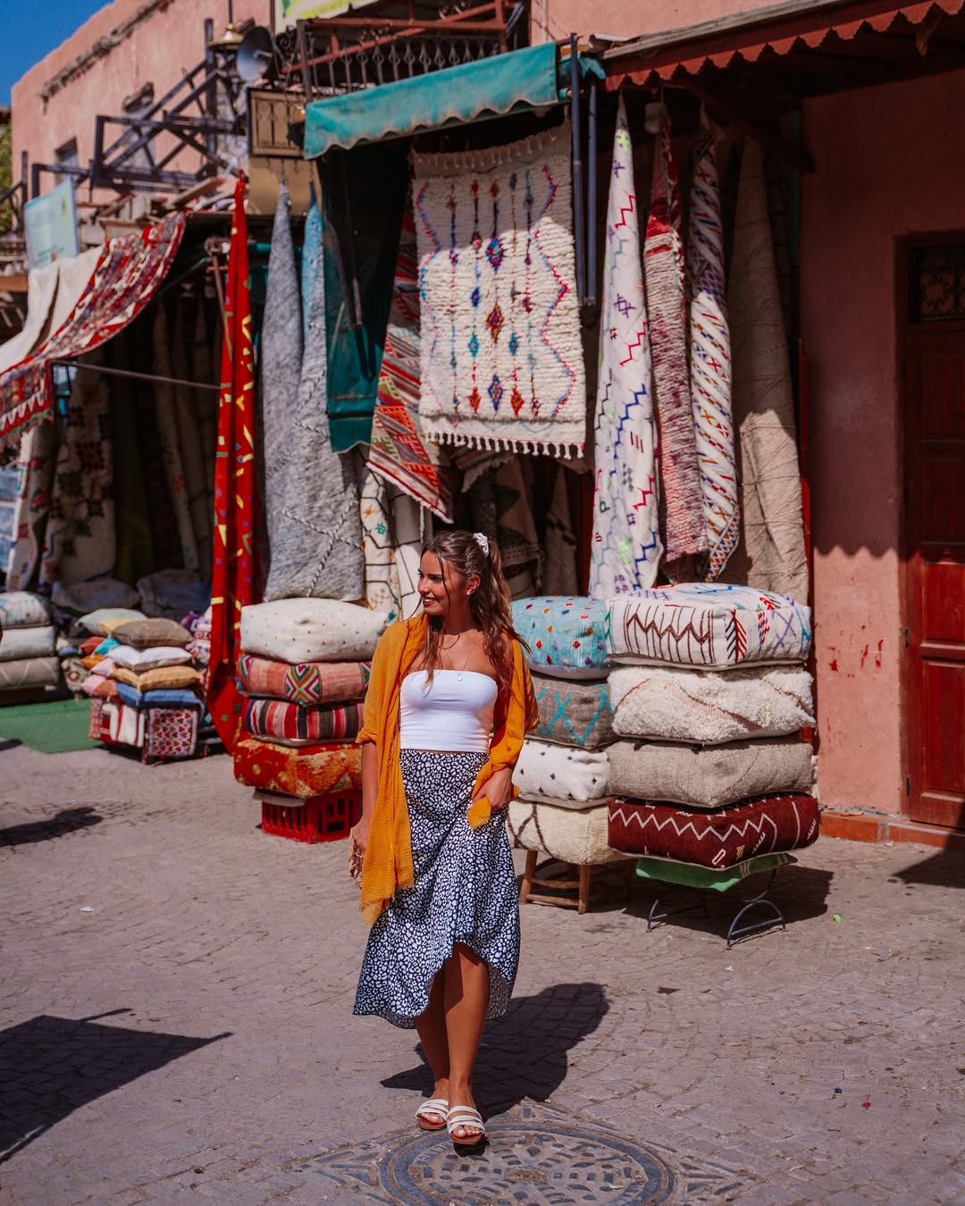 Traveller walking past hanging Berber rugs and stacked floor cushions in a Marrakech rug souk