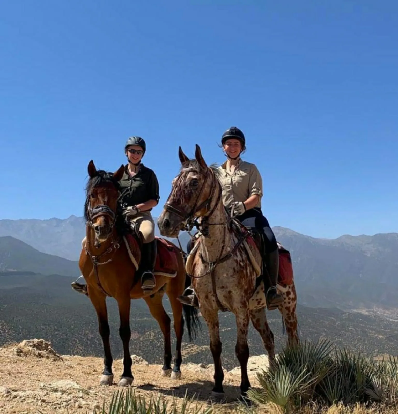 Dos jinetes sonrientes a caballo en un mirador con la cordillera del Alto Atlas de fondo