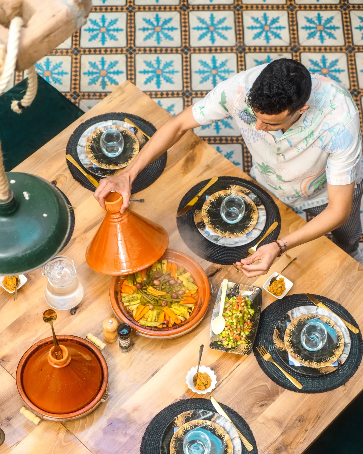 A Moroccan host lifting the lid of a vegetable tagine at a riad dining table