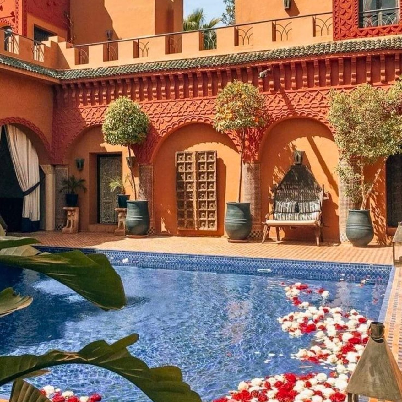 A tiled riad pool scattered with red and white rose petals below carved terracotta arches