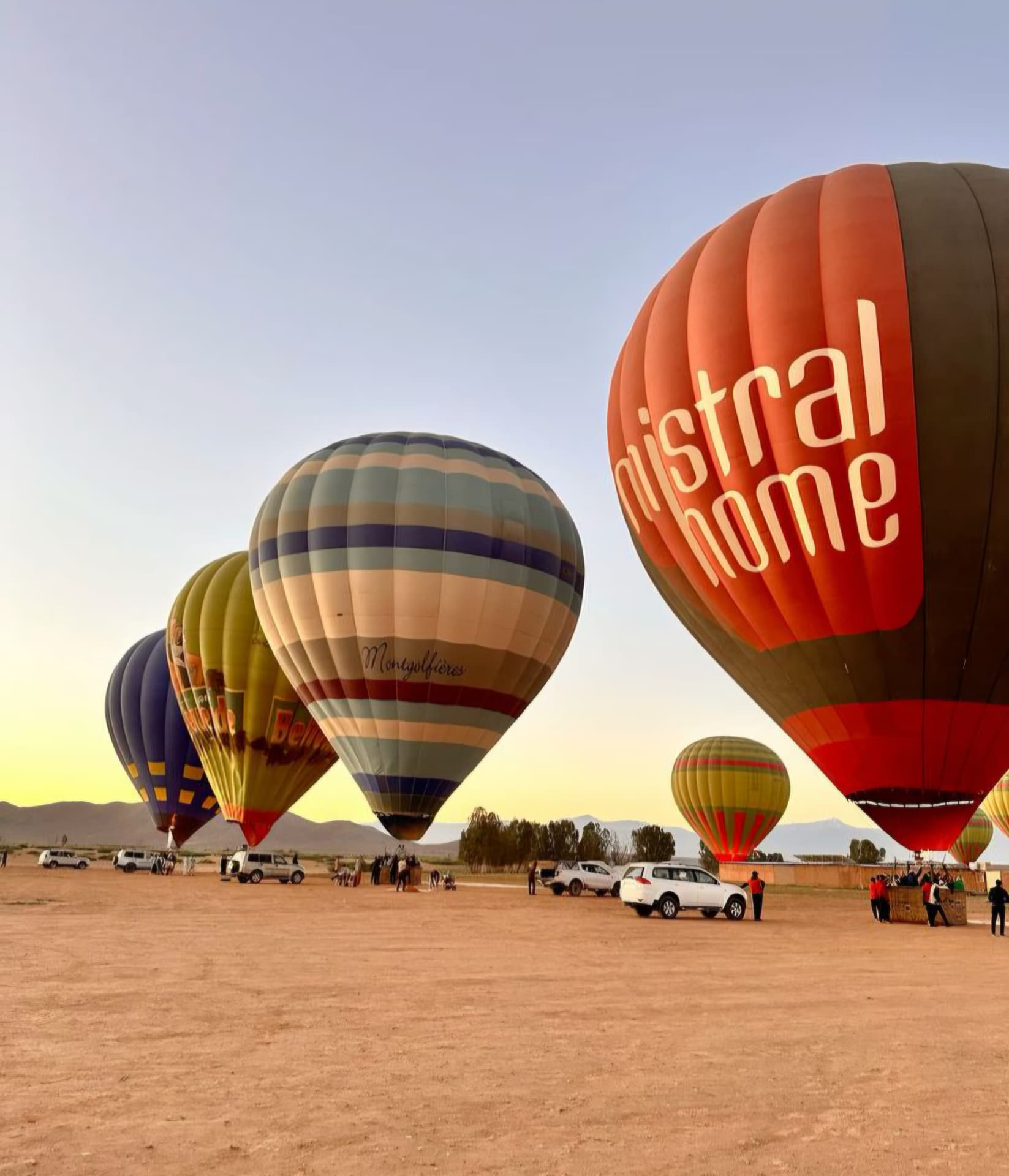 Several hot-air balloons inflating on a dusty launch field at dawn with the Atlas Mountains behind