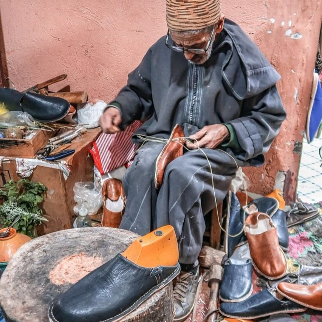 An elderly Moroccan cobbler hand-stitching a leather babouche surrounded by his tools