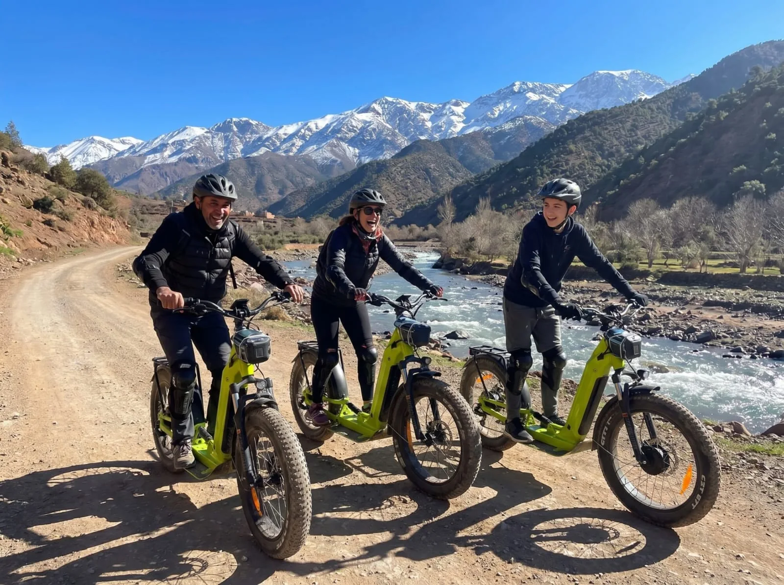 Three riders on off-road e-scooters beside the Ourika river below snow-covered High Atlas peaks