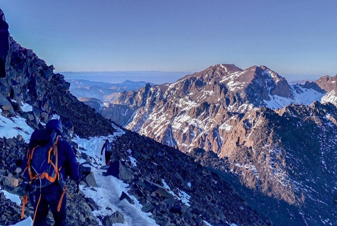 A trekker descending a snowy slope at first light with lit Toubkal peaks across the valley