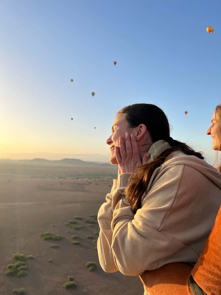 Two passengers smiling in a balloon basket at sunrise, other balloons in the sky