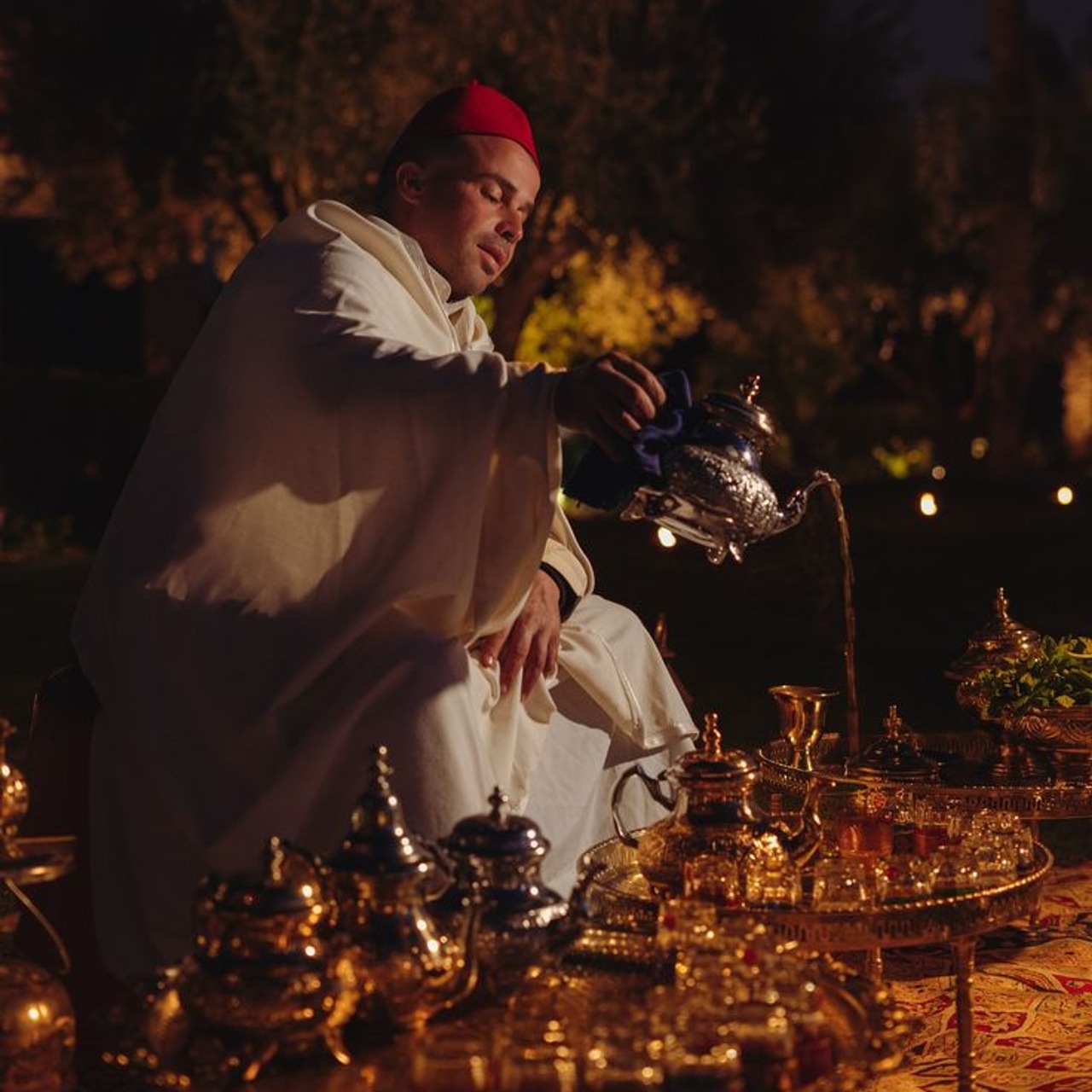 A host in white robes and a red fez pouring tea by candlelight beside a tray of silver teapots in a garden at night