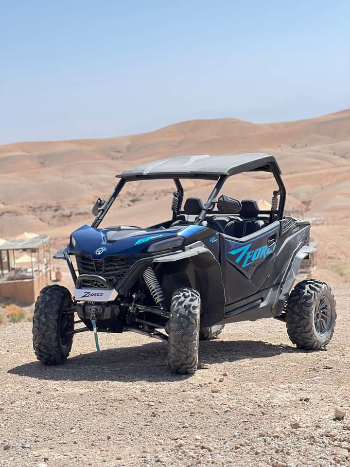 A blue two-seater buggy parked on a gravel ridge in the Agafay desert beside a camp parasol, with the hills behind