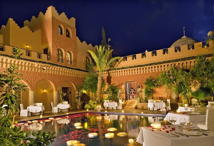 Floodlit kasbah courtyard at Kasbah Tamadot in the evening, with a lit pool, arched terraces and tables laid for dinner