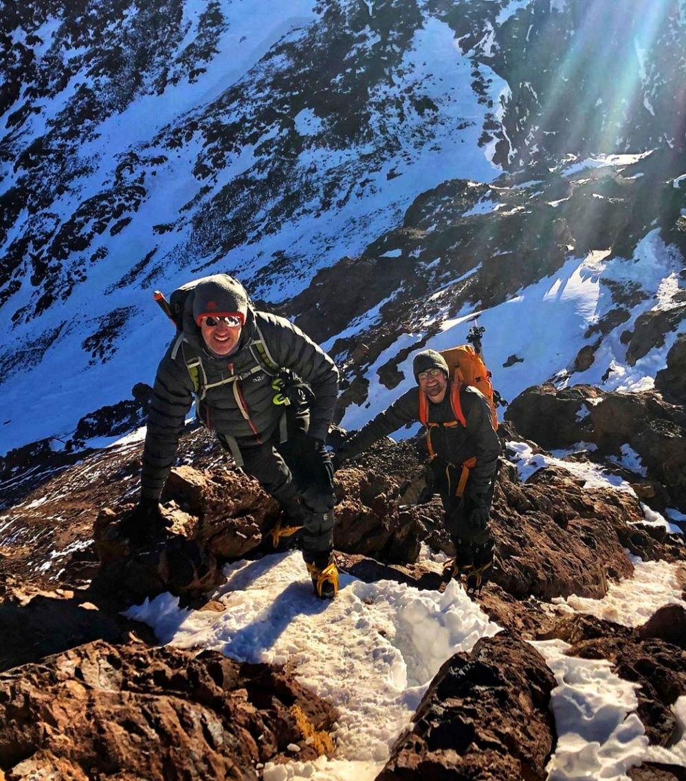 Two climbers in crampons and winter kit smiling as they scramble up rock and snow near the summit