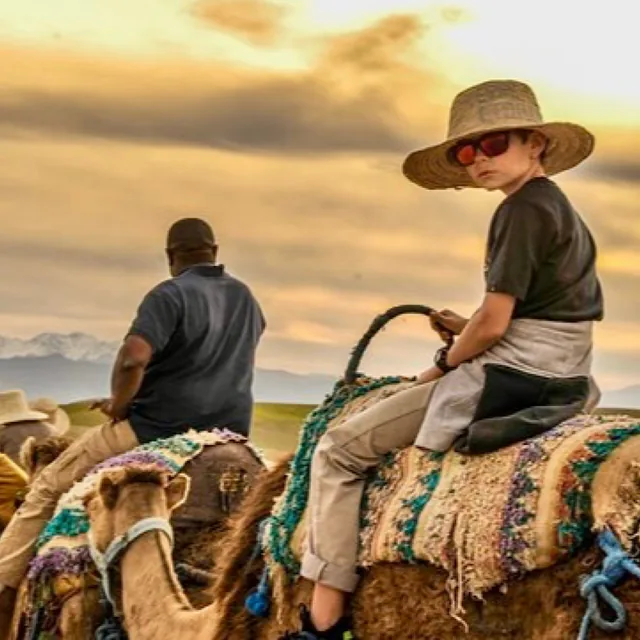A boy in a straw hat and sunglasses riding a camel at sunset with the snow-capped Atlas mountains behind