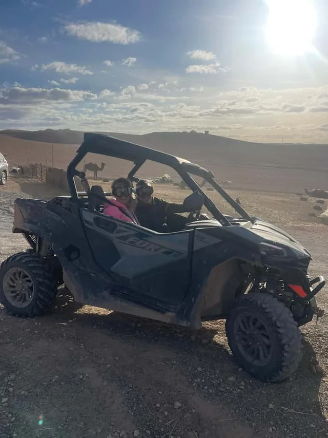 Due persone con casco e occhiali su un buggy biposto nel deserto di Agafay vicino a Marrakech, con un cammello sullo sfondo