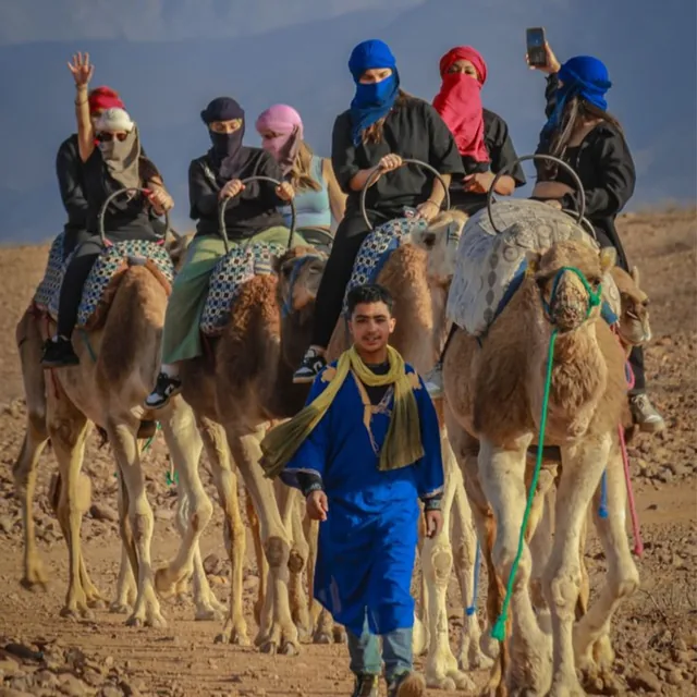 A young camel guide in a blue robe leading riders wearing colourful turbans along a stony desert track
