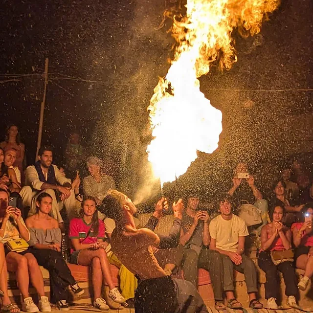 A fire breather sending a tall flame into the night sky in front of a seated audience at a desert camp