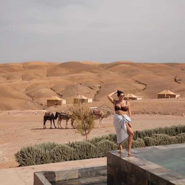 A guest by the pool of a desert camp with tents and saddled camels on the hills behind