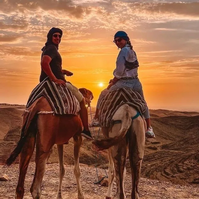 Two riders on camels turning back towards the camera as the sun sets over the stony Agafay desert