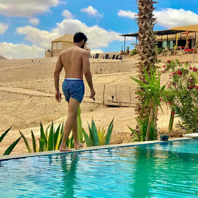 A guest walking along the edge of a turquoise pool at a desert camp with tented lodges and stony hills behind