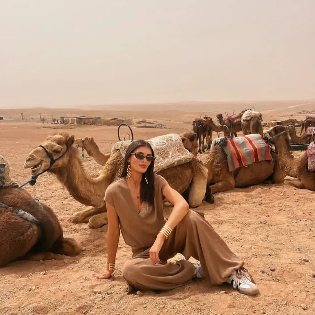 A traveller sitting on the sand beside resting camels with woven saddle blankets in the Agafay desert