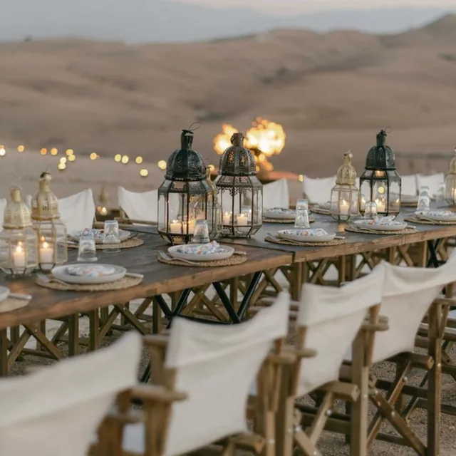 A long wooden dinner table set with lanterns and candles facing the Agafay hills at dusk