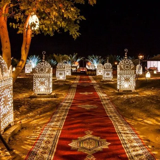 A carpeted walkway lined with glowing carved lanterns leading into a desert camp at night