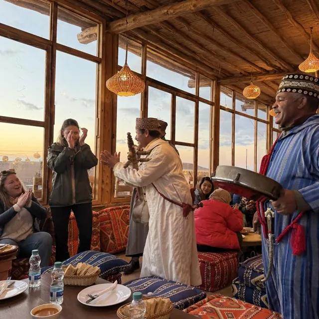 Musicians in traditional dress playing a frame drum for clapping guests in a glass-walled desert dining room at sunset