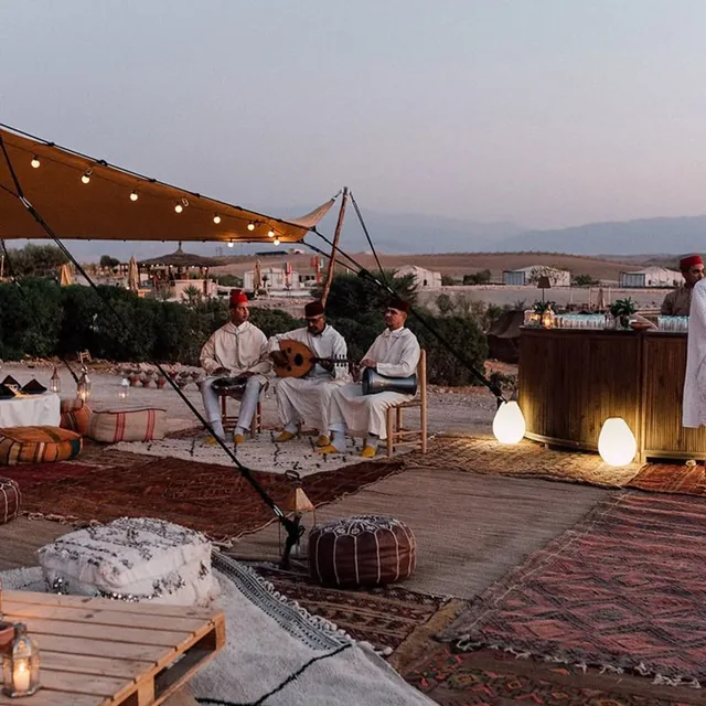 Three Moroccan musicians in white robes playing oud and drums in a rug-covered camp lounge at dusk