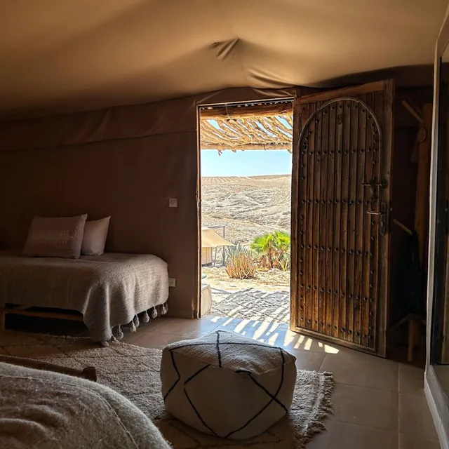 The inside of a canvas desert lodge with a low bed and an open carved wooden door onto the hills
