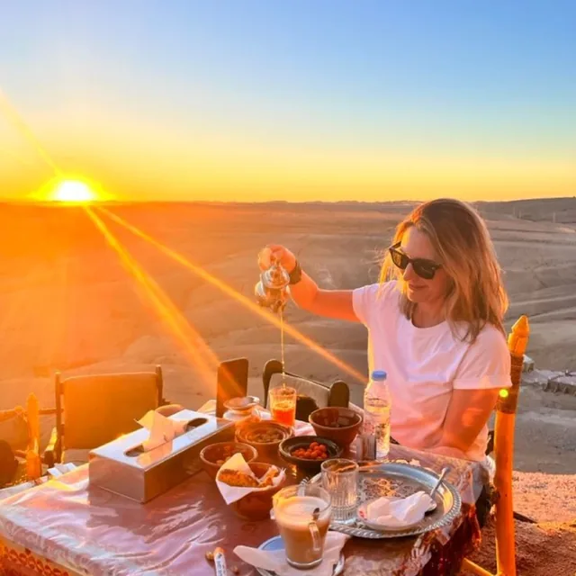 A guest pouring mint tea from a silver pot at a table set with pastries as the sun sets over the desert