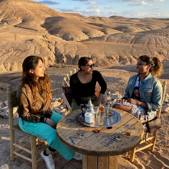 Three friends sharing a tray of mint tea at a wooden table overlooking the Agafay desert hills