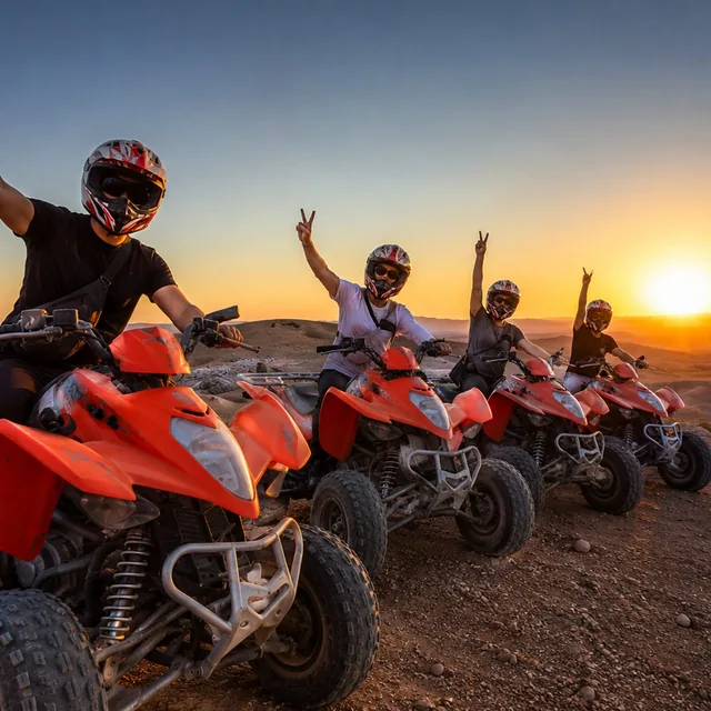 Four helmeted riders on orange quad bikes raising their hands as the sun sets over the hills
