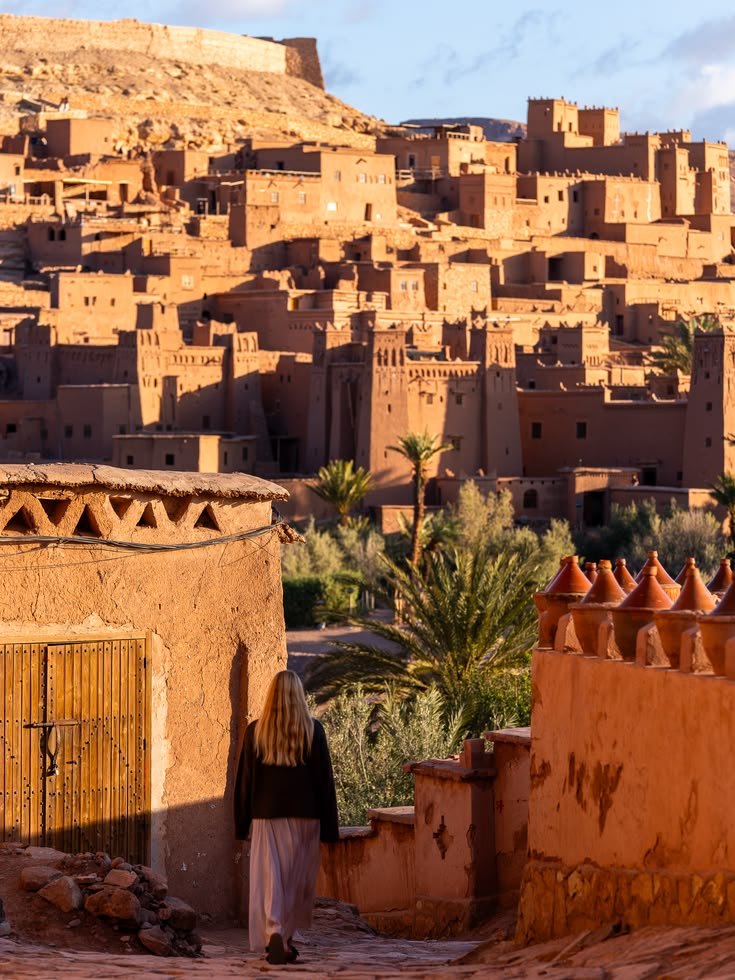 A traveller walking an earthen lane towards the terraced towers of Aït Ben Haddou in warm late-day light, with clay crenellations along a wall