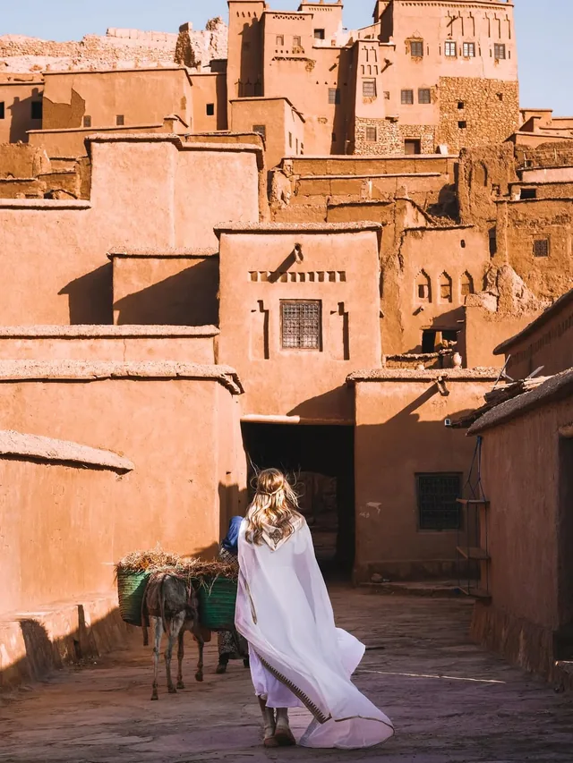 A traveller walking a sunlit earthen lane inside the Aït Ben Haddou ksar as a loaded donkey passes