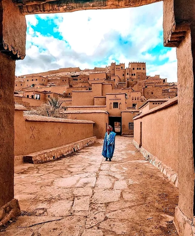 A villager in a blue robe walking a stone lane below the earthen houses of Aït Ben Haddou