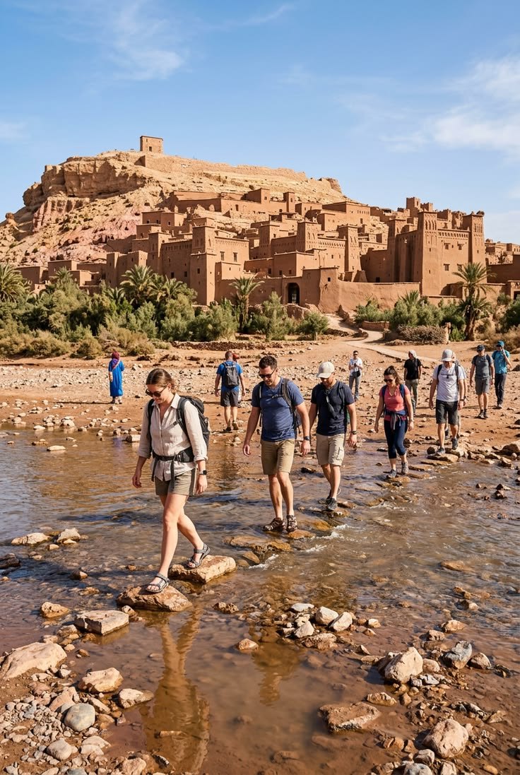 A small group of walkers crossing a shallow river on stepping stones below the earthen ksar of Aït Ben Haddou