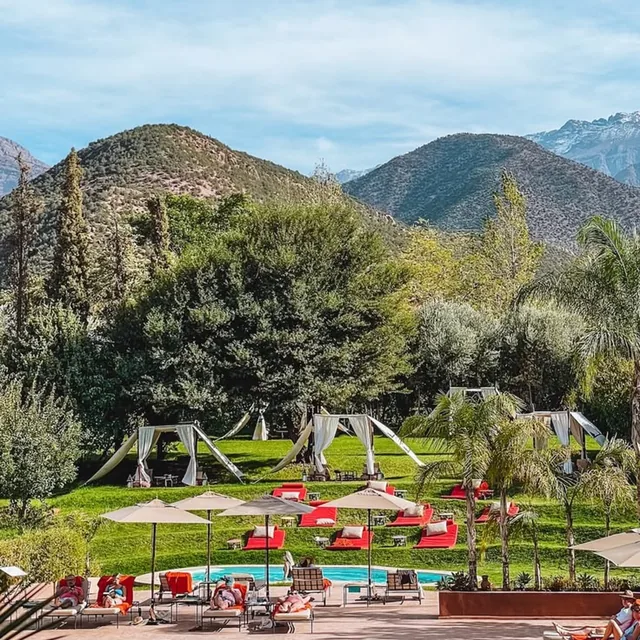 A garden pool with loungers and shaded tents below the green foothills and snow-capped High Atlas
