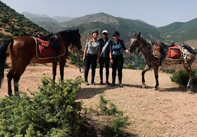 Dos jinetes y un guía con sus caballos en un sendero de montaña con las verdes laderas del Atlas al fondo