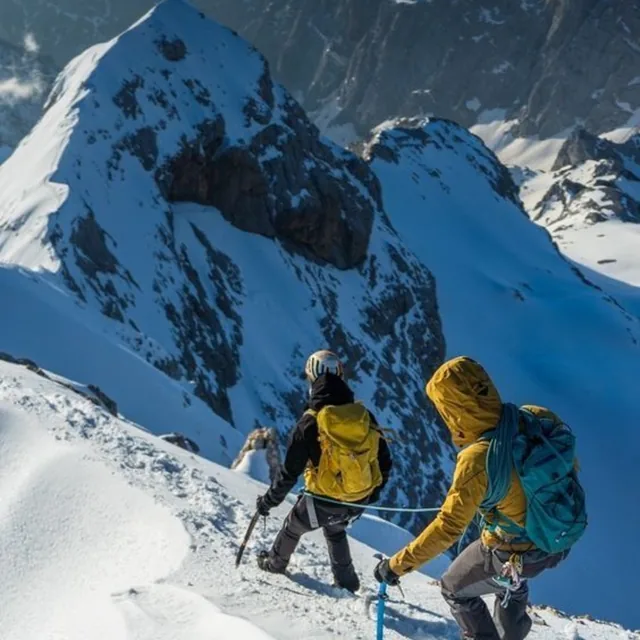 Two roped mountaineers in helmets and crampons moving along a snow ridge with a rocky summit behind
