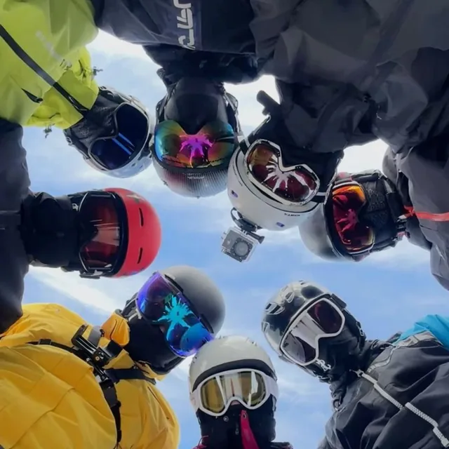 A circle of skiers in helmets and goggles looking down at the camera against a bright sky
