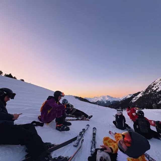 A group of skiers sitting on the snow with their skis beside them as the sky turns orange at sunset